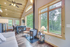 A spacious living room with a large fireplace, a ceiling fan, and a view of the forest through large windows. - Dock, Fire Pit, Games! Spirt Lake Family Retreat (Frederic)