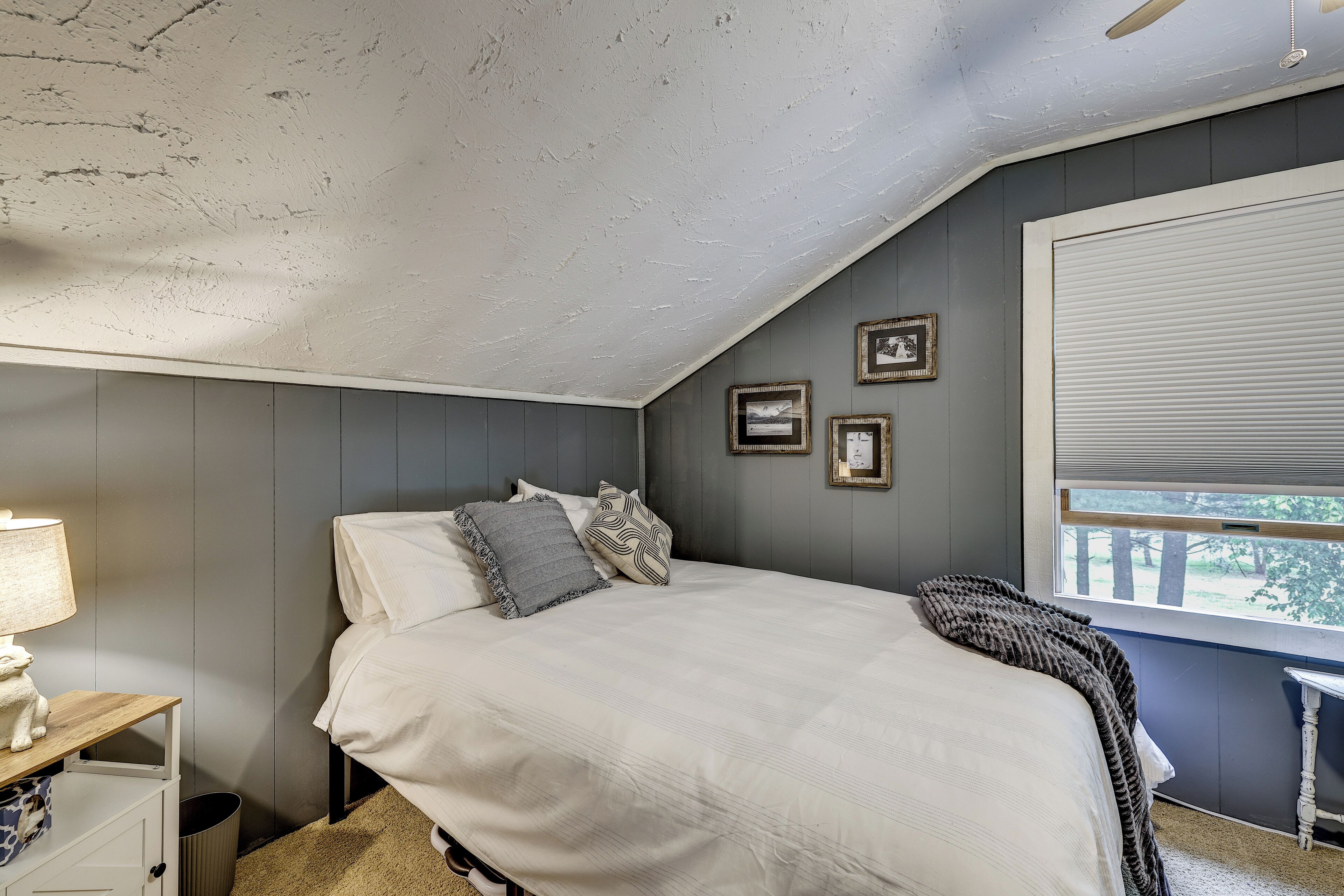 A bedroom with a sloped ceiling, a bed with pillows, a nightstand with a lamp, and framed pictures on the wall.