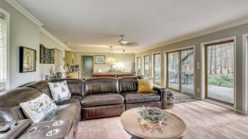 A spacious living room with a large sectional sofa, a glass coffee table, and a view of a wooded area through large windows.