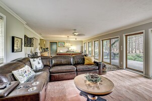 Uma sala de estar ampla com um sofá seccional grande, uma mesa de centro de vidro e vista para uma área arborizada através de grandes janelas.