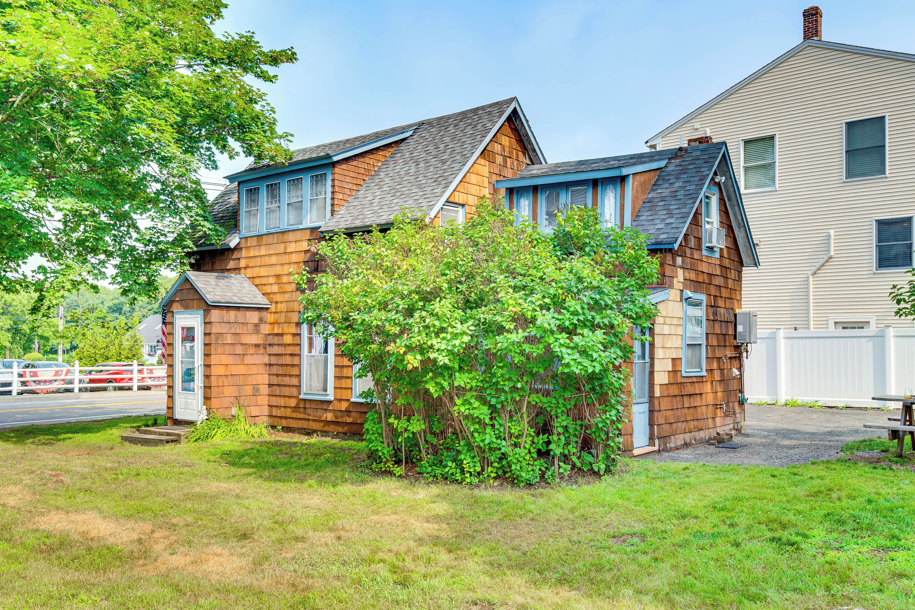 A two-story wooden house with a small garden and a white fence in the background.