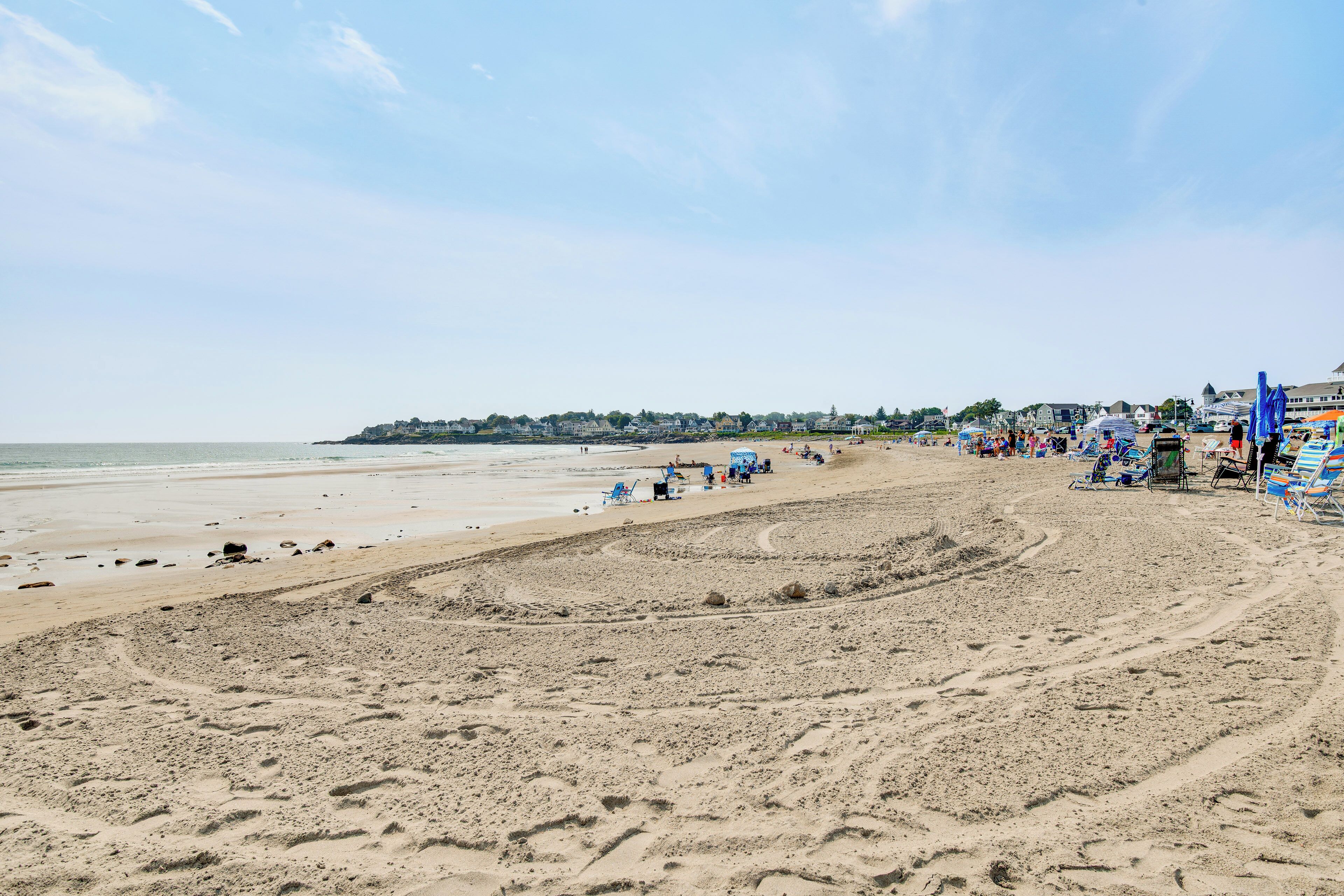 Una playa con arena, sillas de playa, sombrillas y una fila de casas al fondo.
