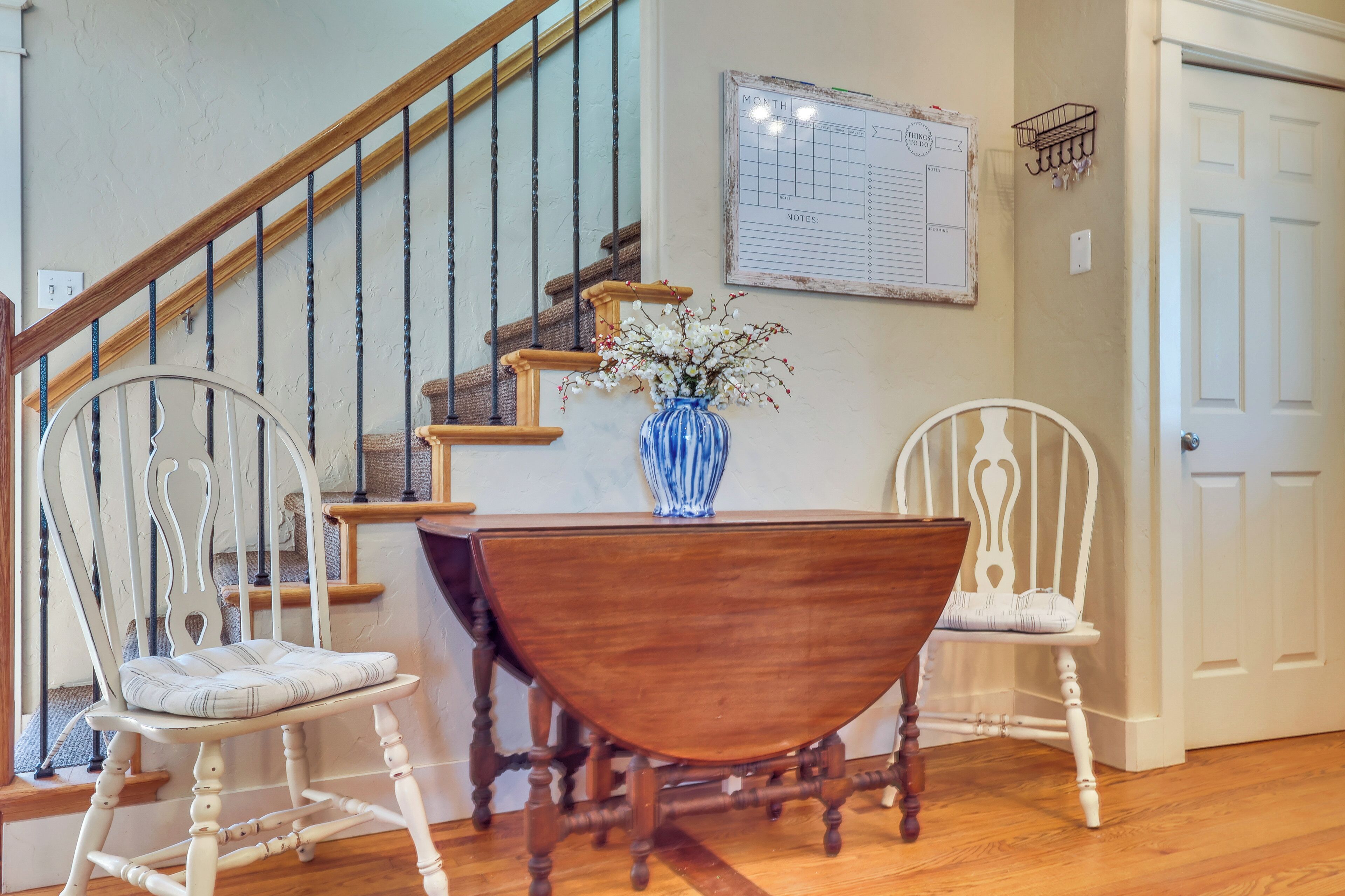 A staircase with a wooden handrail and metal balusters, a dining table with a vase of flowers, and two white upholstered chairs.