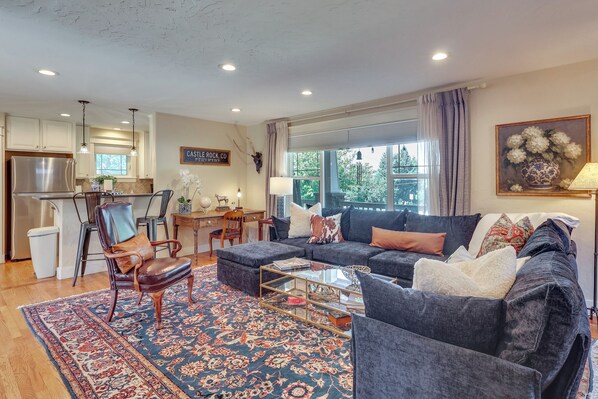 A spacious living room with a sofa, armchairs, a coffee table, and a rug. There is a kitchen area with cabinets and a refrigerator.
