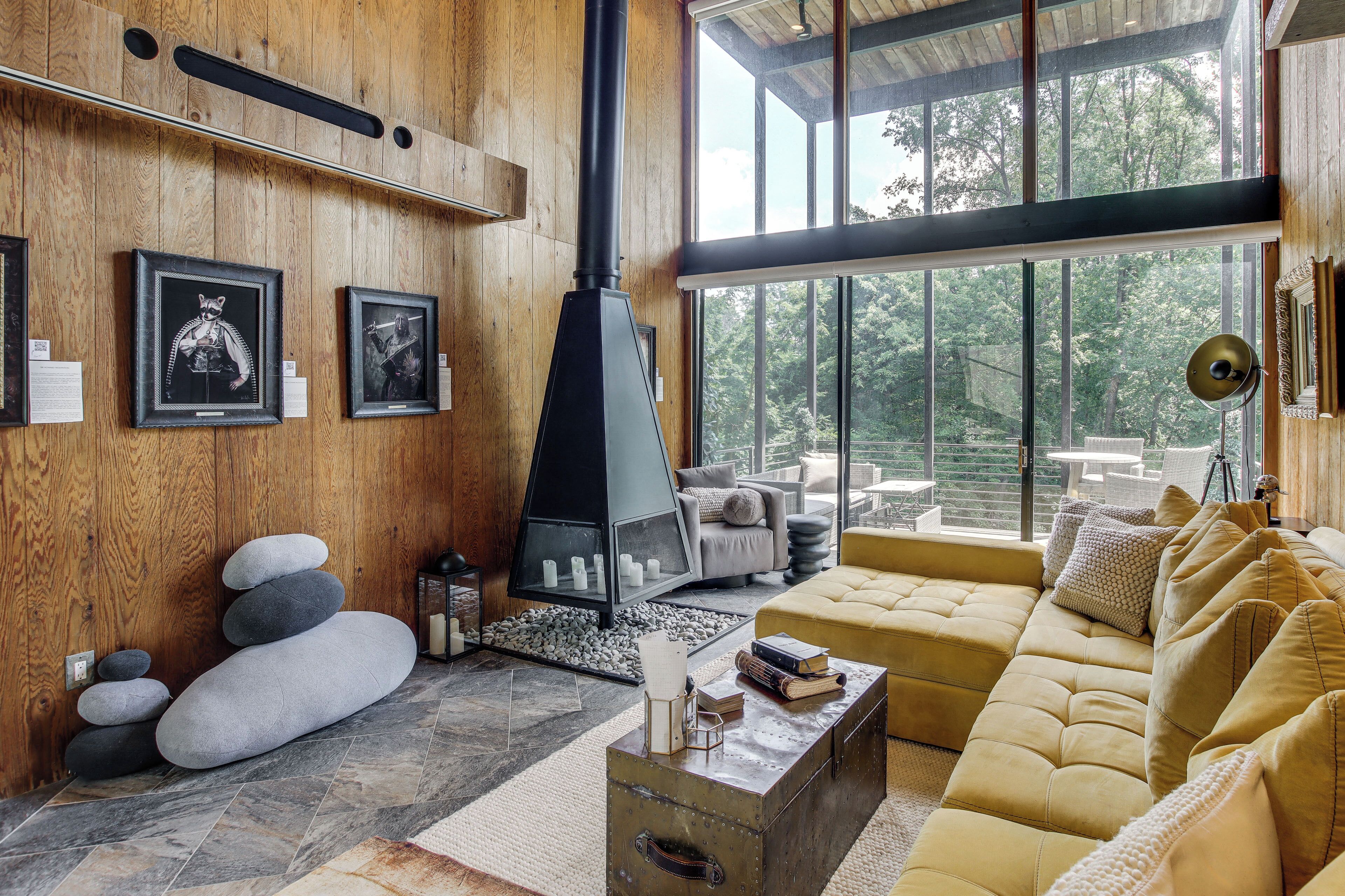 A modern living room with a stone fireplace, wooden walls, and large windows offering a view of the outdoors.
