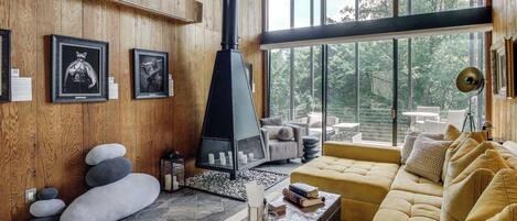 A modern living room with a stone fireplace, wooden walls, and large windows offering a view of the outdoors.