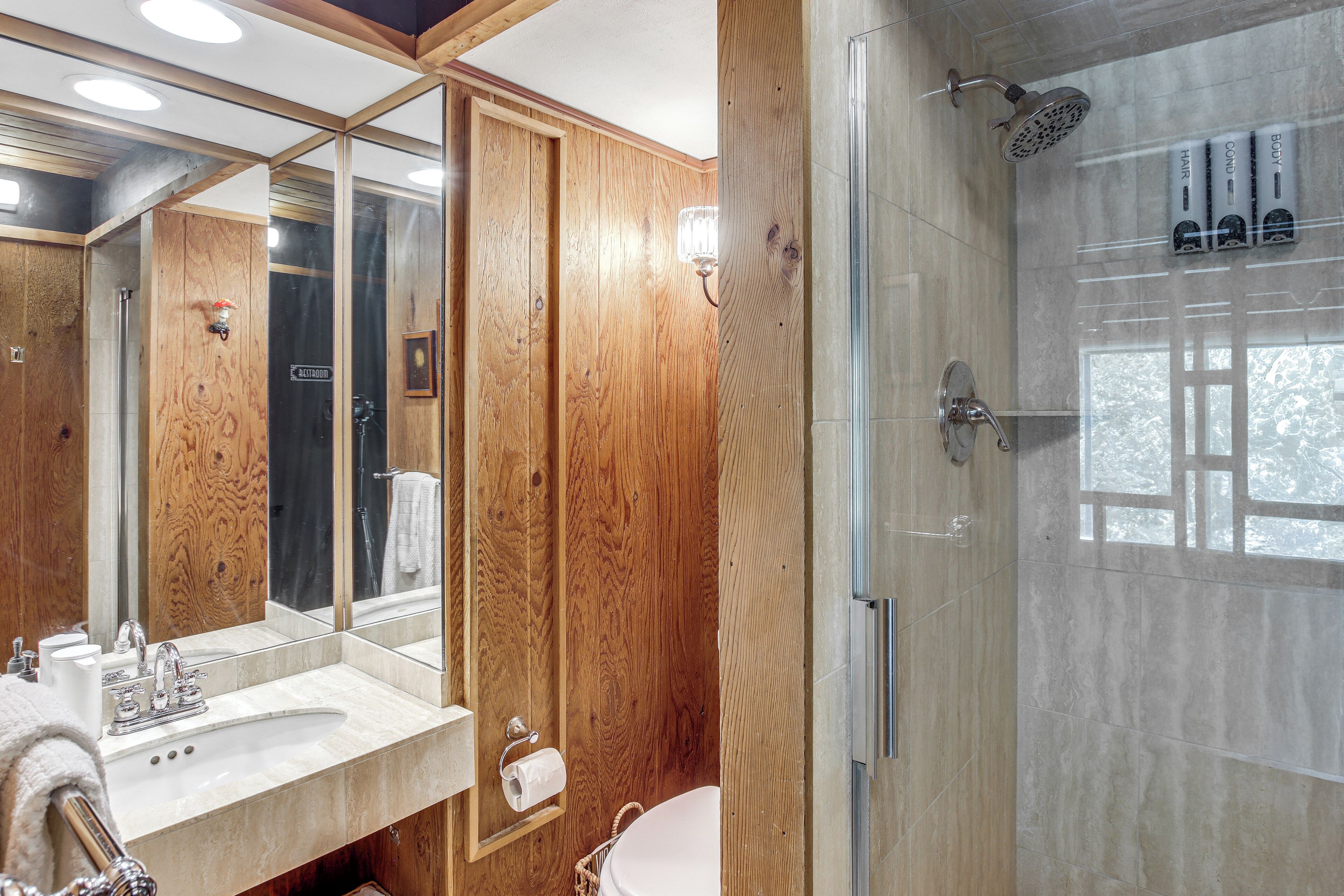 A bathroom with a wooden vanity, a large mirror, a glass shower enclosure, and a toilet paper holder.