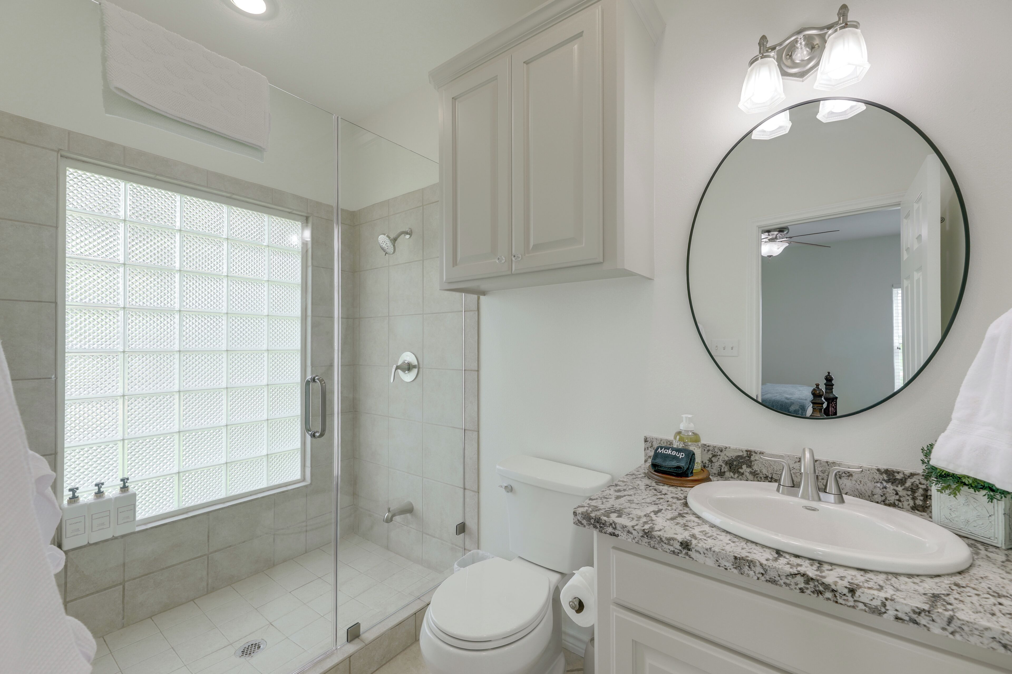 A bathroom with a glass shower enclosure, a toilet, a sink with a marble countertop, and a mirror with lights.