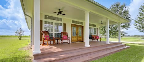 A covered porch with wooden chairs and a bench, a wooden door, and a view of a green landscape.