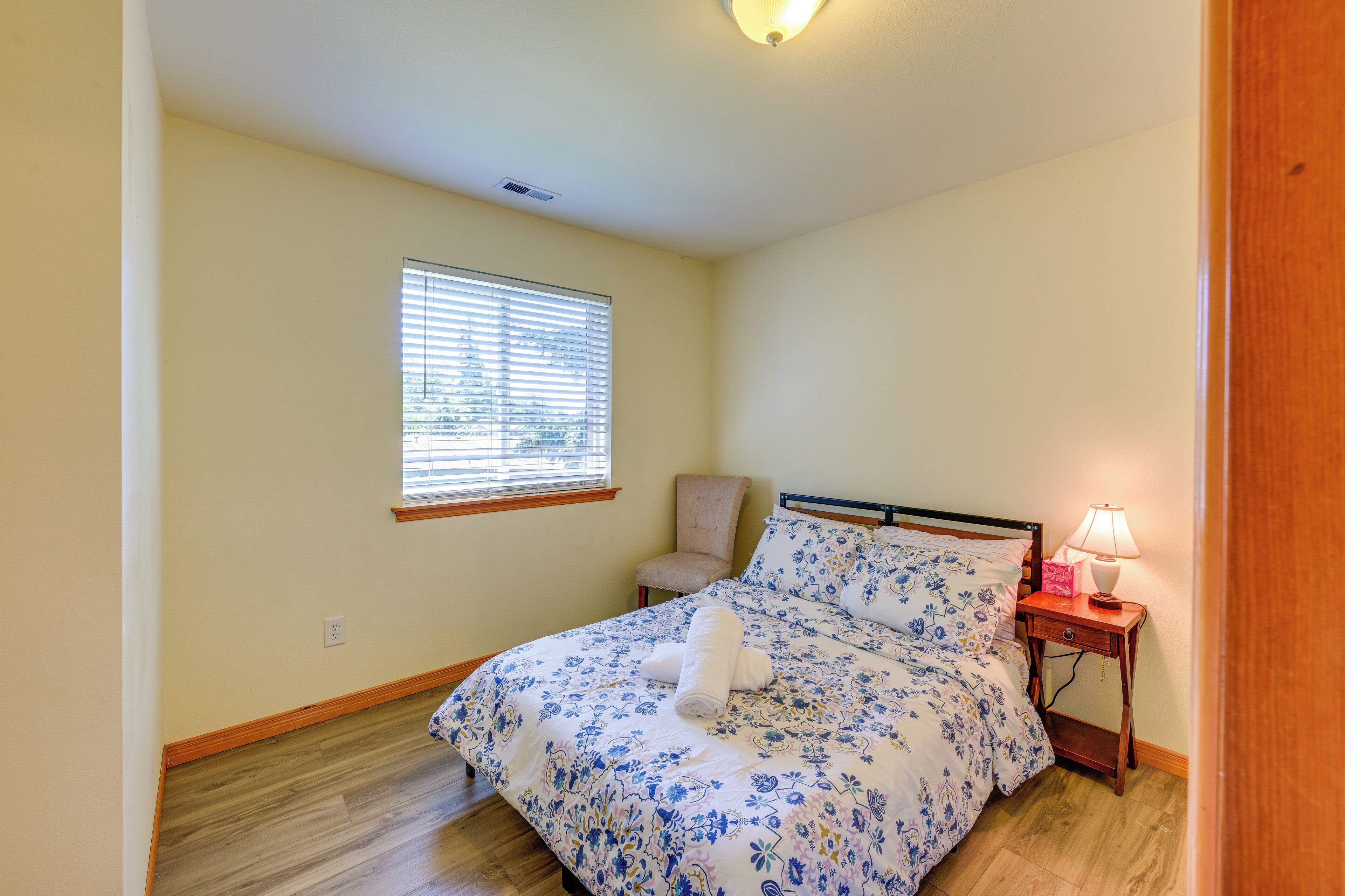 A bedroom with a bed, a chair, a bedside table with a lamp, and a window with blinds.