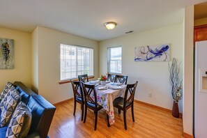 A dining area with a table set for four, wooden chairs, a floral tablecloth, and two windows with blinds.