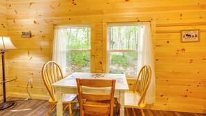 Interno di una baita di legno con un tavolo e sedie, due finestre con tende, una lampada e un cartello appeso al muro.