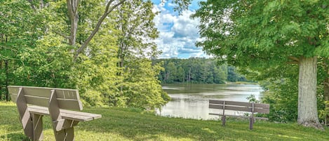 Un parque con dos bancos de madera, una zona verde y un lago rodeado de árboles.