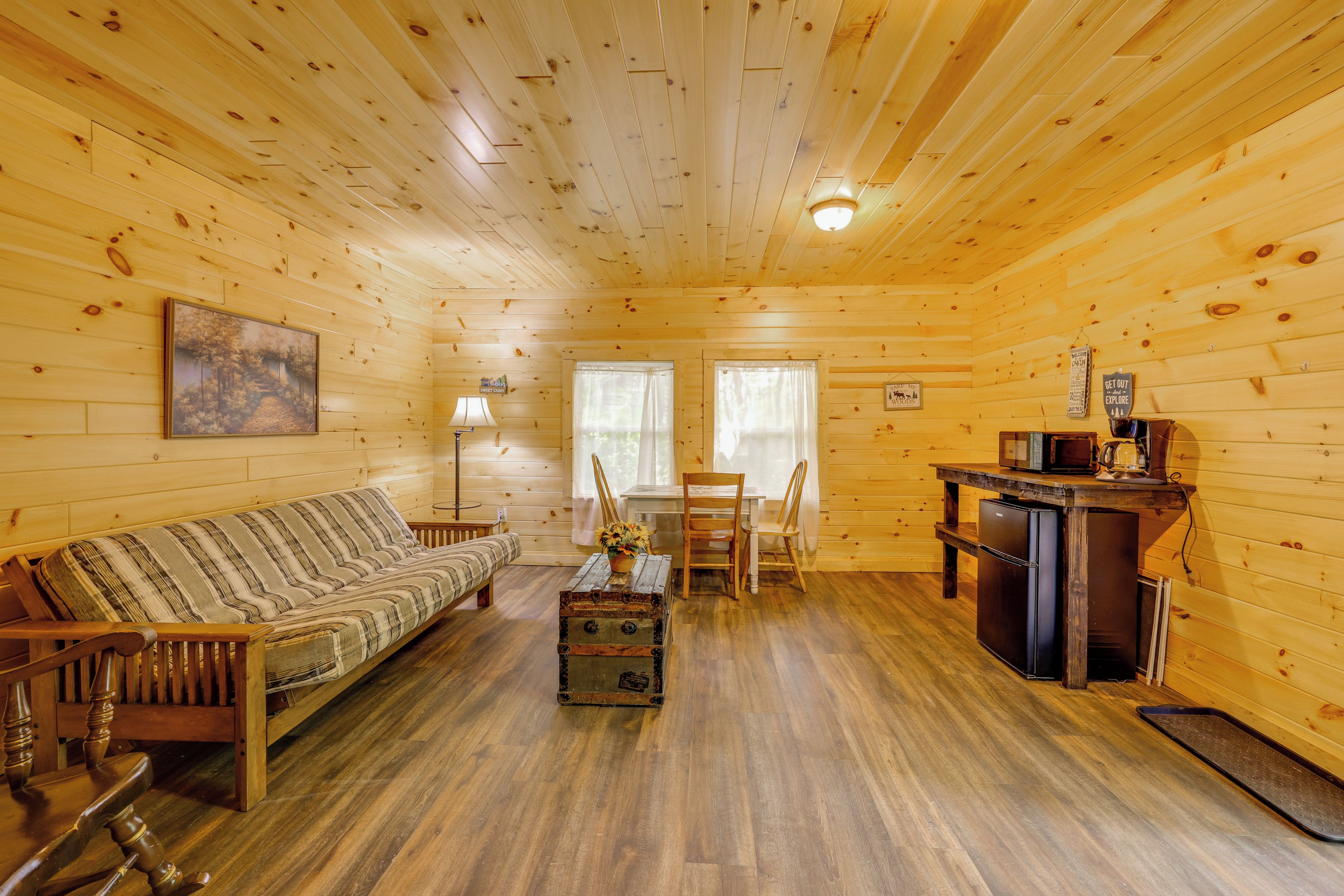 A cabin interior with a wooden floor, a sofa, a dining table with chairs, a small kitchenette, and a painting on the wall.