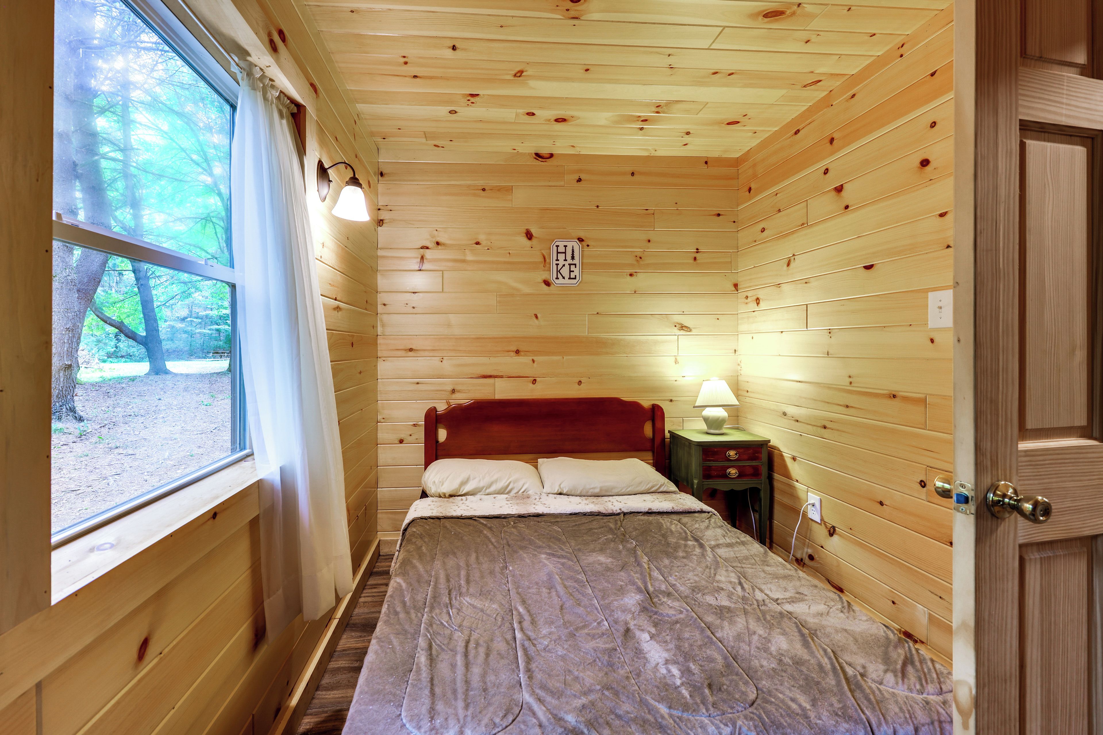 A wooden cabin bedroom with a bed, a nightstand, a lamp, and a window overlooking a forest.