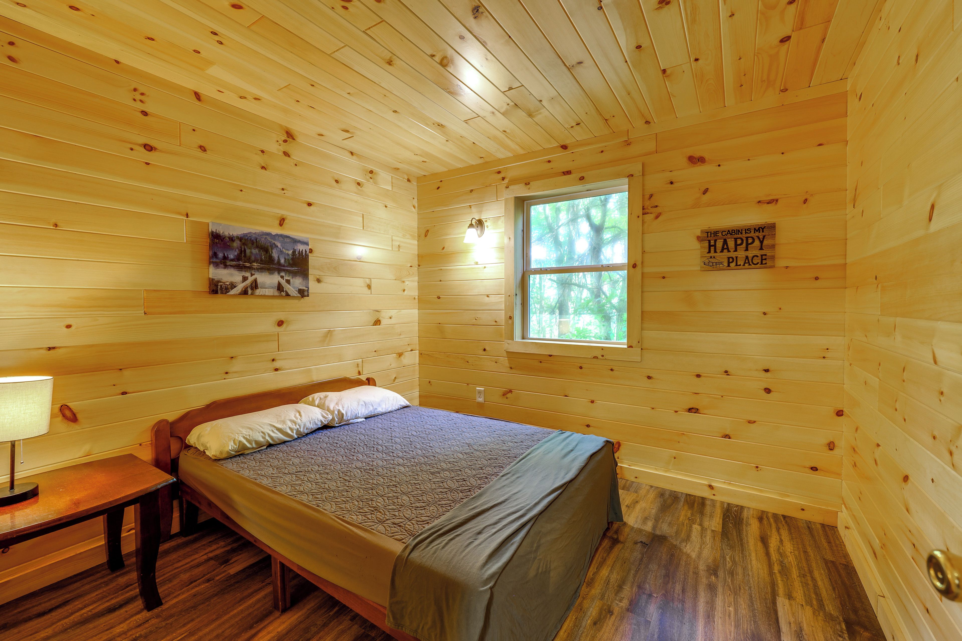 A wooden cabin bedroom with a bed, a nightstand, a lamp, a painting, and a window.
