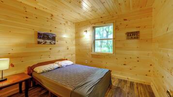 A wooden cabin bedroom with a bed, a nightstand, a lamp, a painting, and a window.
