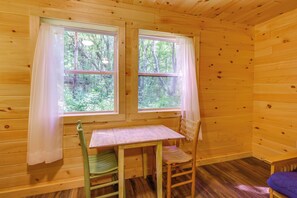 A wooden cabin interior with a table and two chairs, two windows with curtains, and a view of the forest. - Fun-filled Getaway at Letchworth Valley Resort (Bliss)