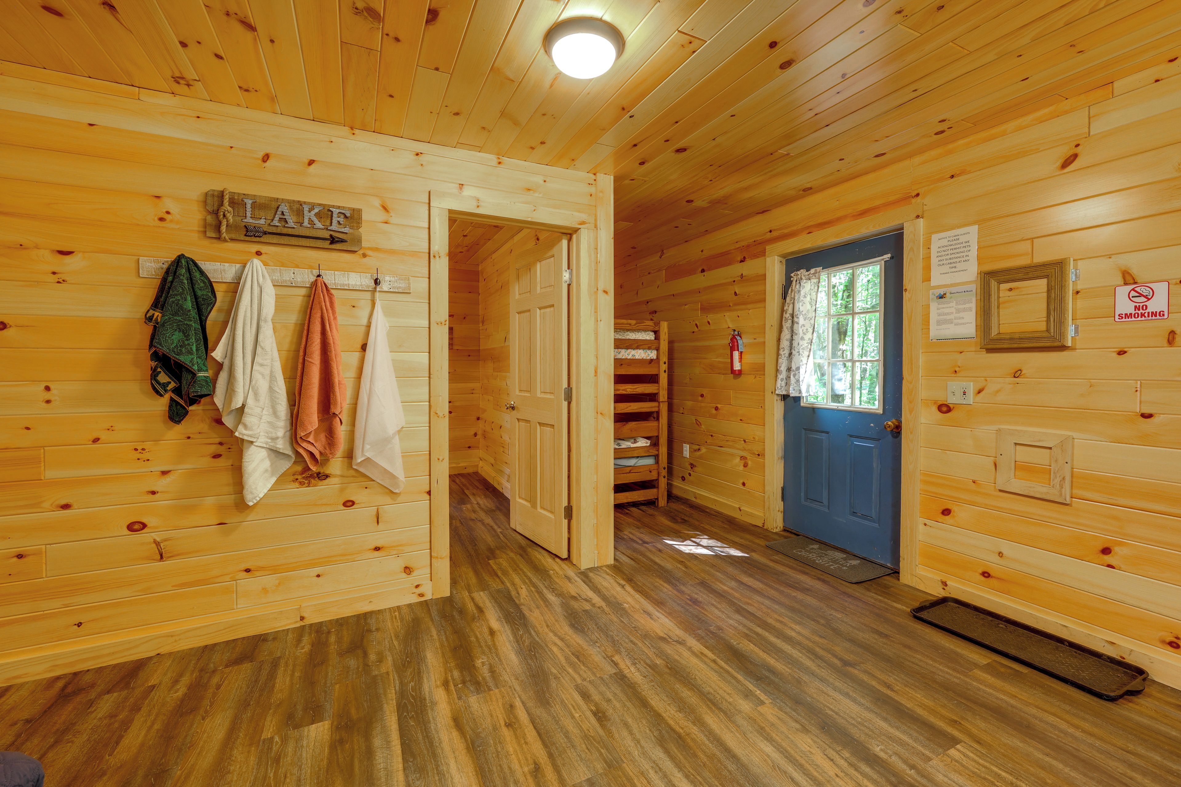 A wooden cabin interior with a hallway leading to a door, a sign indicating 'LAKE', and a 'NO SMOKING' sign.