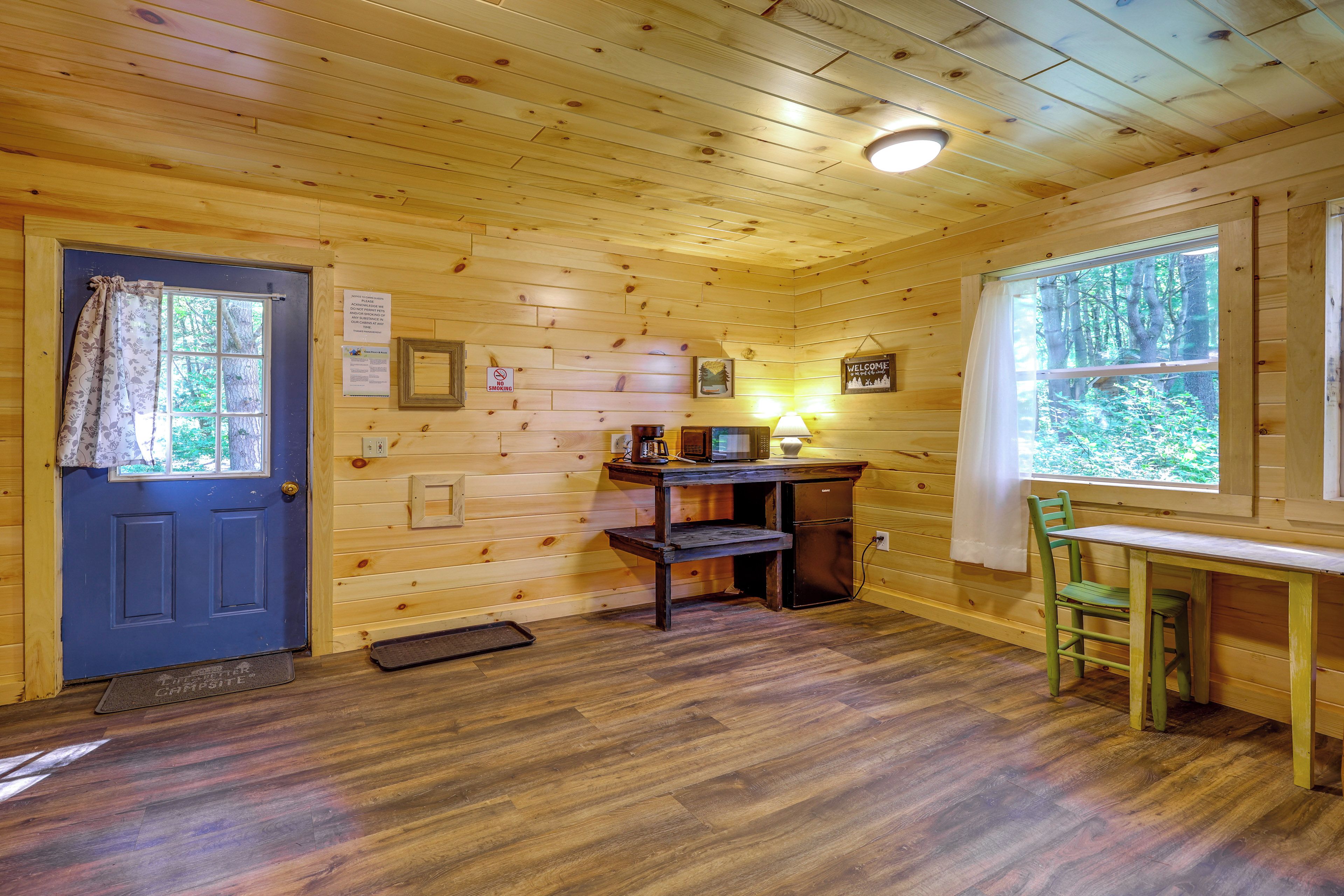 A cabin interior with a wooden door, a desk with a computer, a chair, and a window with a view of trees.