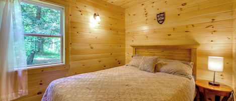 A wooden cabin bedroom with a bed, a nightstand, a lamp, a window with curtains, and a sign on the wall.