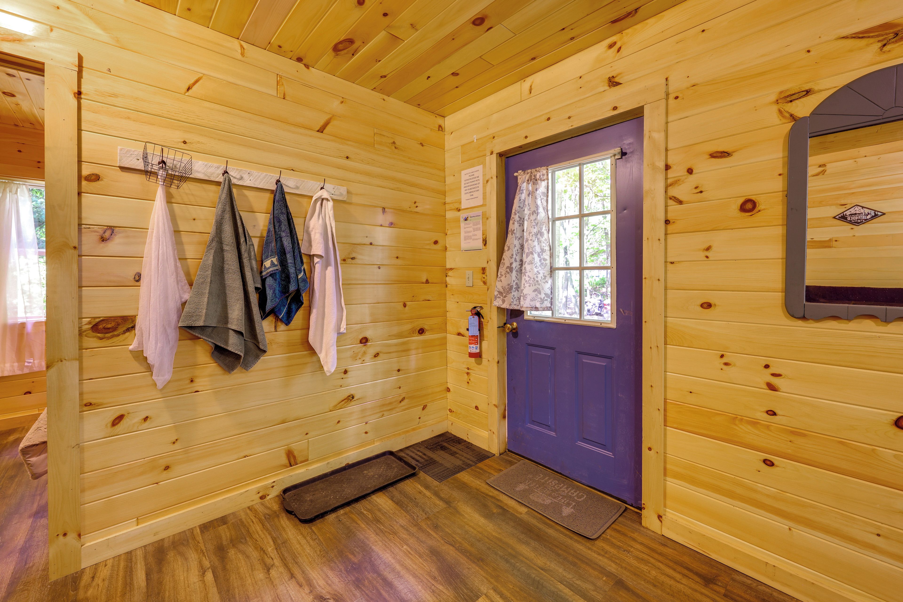 A wooden cabin interior with a purple door, a towel rack with several towels, a fire extinguisher, and a window with a curtain.