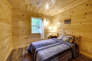 A wooden cabin bedroom with a bed, a nightstand, a lamp, a window, and a welcome sign. - Fish, Slide & Unwind: Letchworth Valley Cabin! (Bliss)