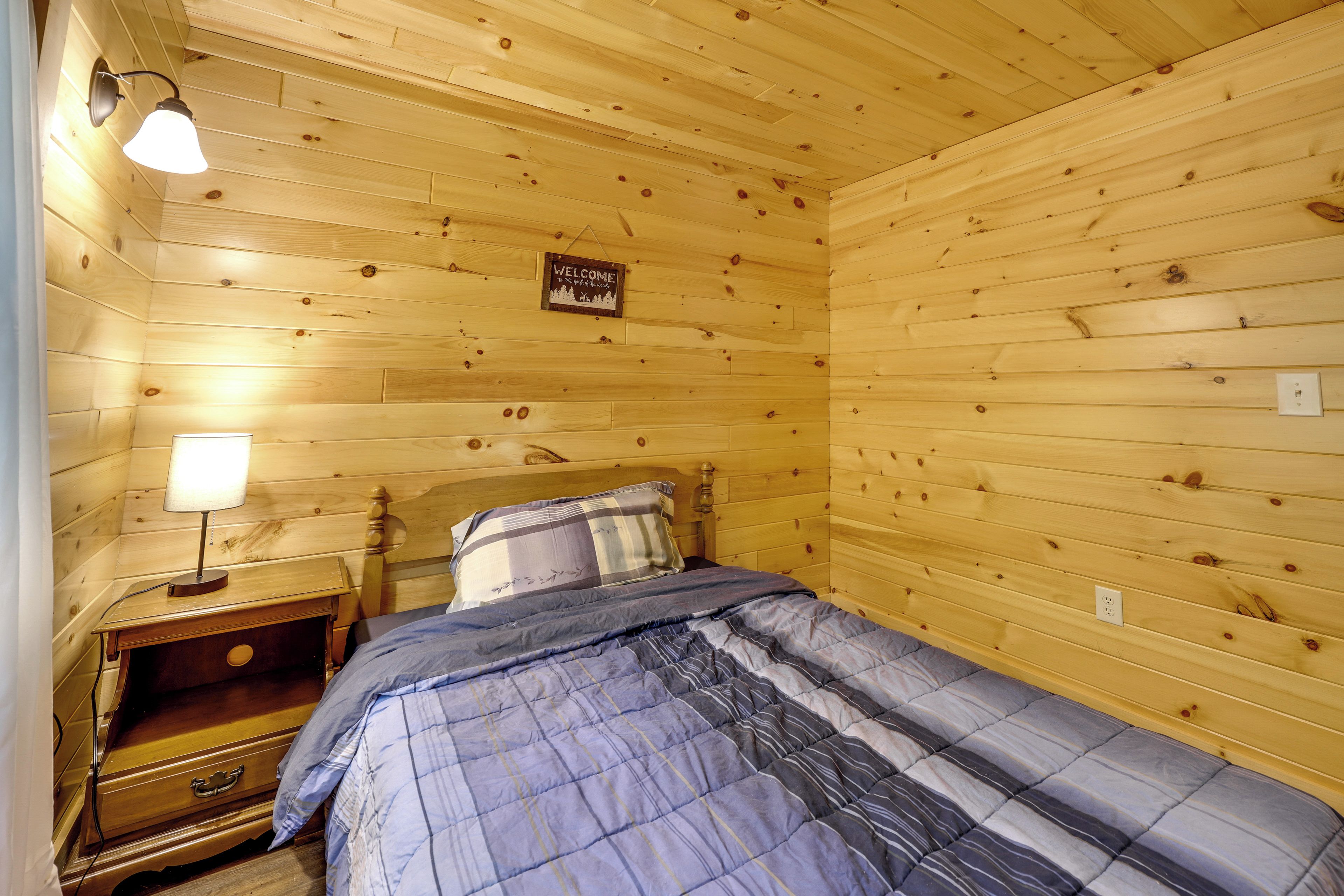 A wooden room with a bed, a nightstand, a lamp, and a welcome sign.