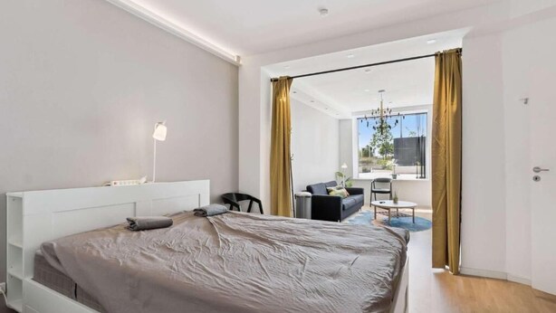 Ein modernes Schlafzimmer mit einem großen Bett, einem Nachttisch, einem Schreibtisch und einem Sessel. Ein Fenster mit Vorhängen bietet Blick ins Freie.