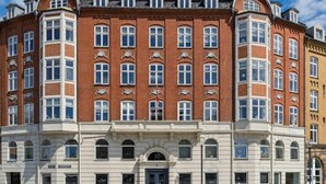 Ein mehrstöckiges Gebäude mit einer symmetrischen Fassade, ein zentraler Eingang mit einer dunkel gefärbten Tür, zahlreiche Fenster in jeder Etage und Zierteile über den Fenstern.