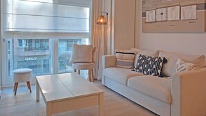 A modern living room with a sofa, a coffee table, and a chair. There is a large window with blinds, a lamp, and a wall-mounted artwork. - Coastal Family Escape in Knokke (Knokke-Heist)
