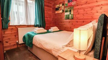 Habitación de cabaña de madera con cama, mesita de noche, lámpara y ventana con cortinas.