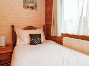 Ein Zimmer mit Holzwänden, einem Bett, einem Nachttisch, einer Lampe und einem mit Vorhängen drapierten Fenster.