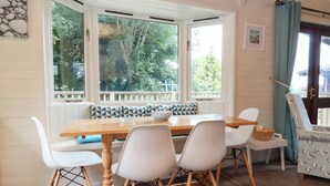 A dining area with a wooden table and white chairs, a bay window with a view of trees, and a patterned cushion on the bench. - Seagull Lodge (Ilfracombe)