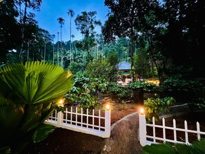 A pathway leading to a building surrounded by lush greenery and a white fence. - Nexgo Spaces Coffee Tales  (Munnar)