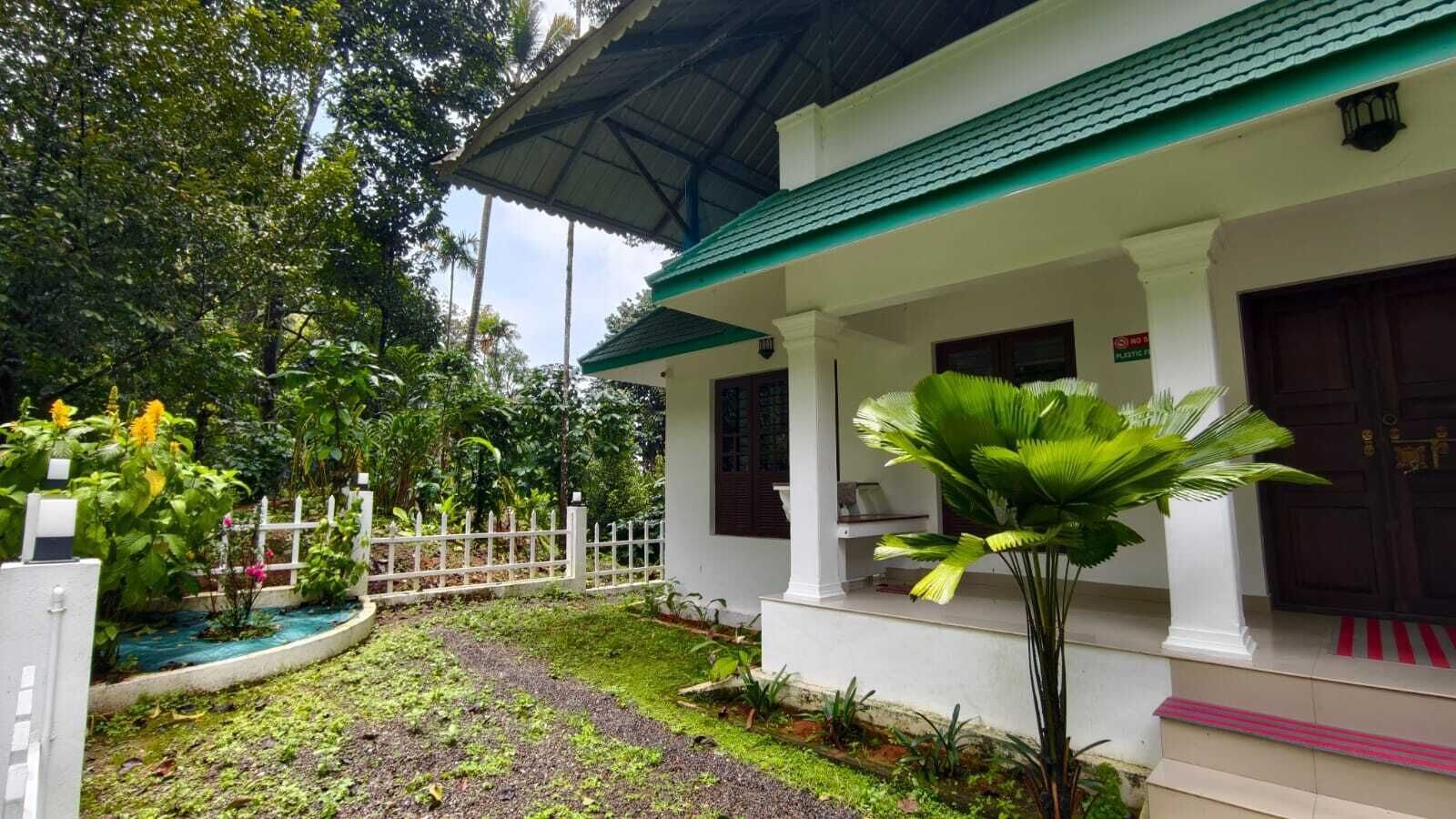 A white building with a green roof, a white fence, and a garden with tropical plants.