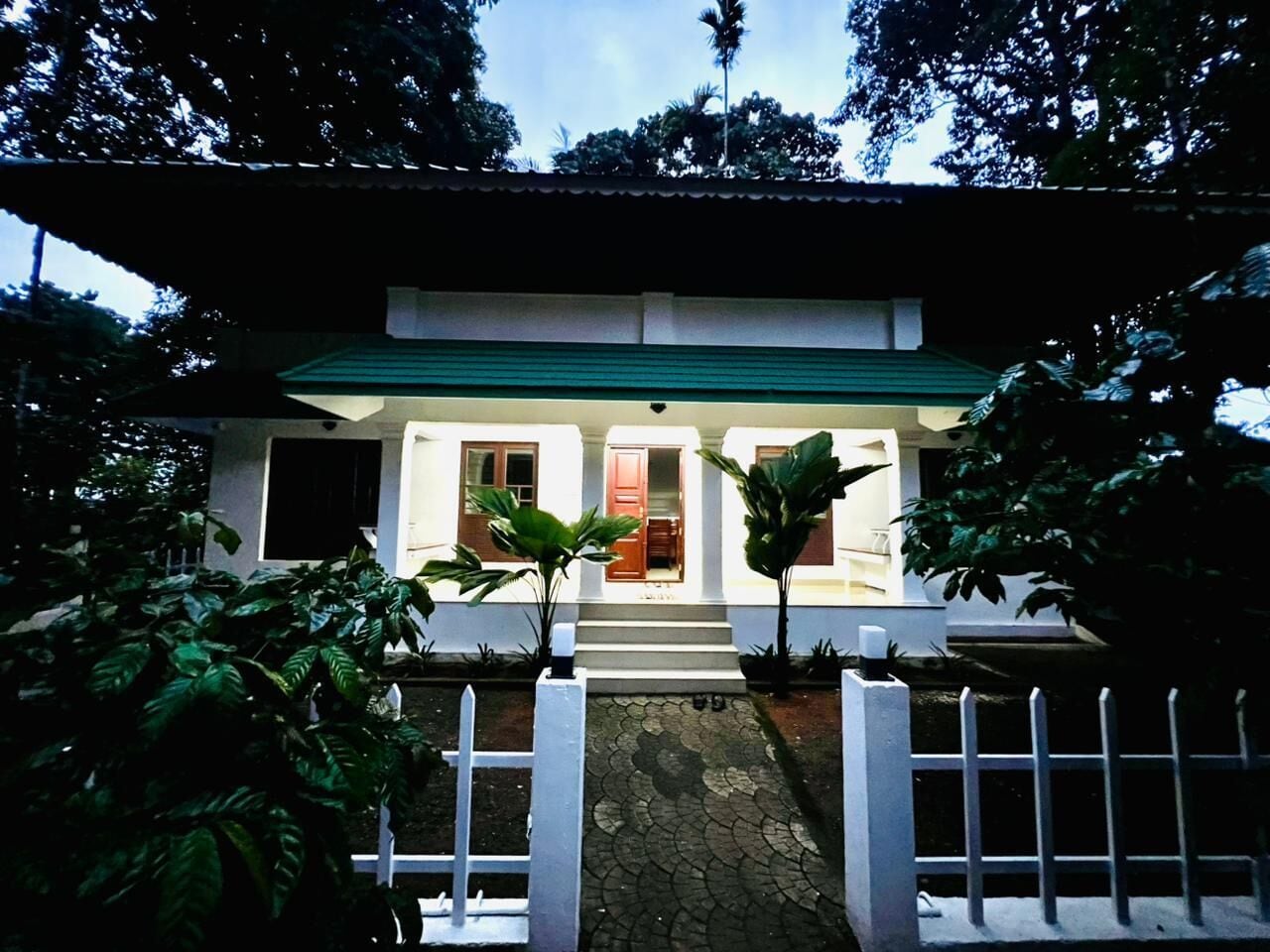 A building with a green roof, white walls, and a white fence in front. There are steps leading to the entrance, and potted plants are placed symmetrically on either side.