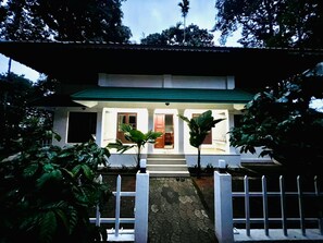 A building with a green roof, white walls, and a white fence in front. There are steps leading to the entrance, and potted plants are placed symmetrically on either side. - Nexgo Spaces Coffee Tales  (Munnar)