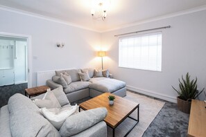 A modern living room with a sectional sofa, a wooden coffee table, a floor lamp, and a large window with blinds. - Bayview Apartment by Guestz (Morecambe)
