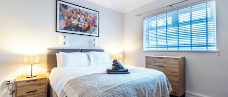 Un dormitorio con cama, mesitas de noche, un tablero auxiliar y un cuadro en la pared, además de una ventana con persianas.
