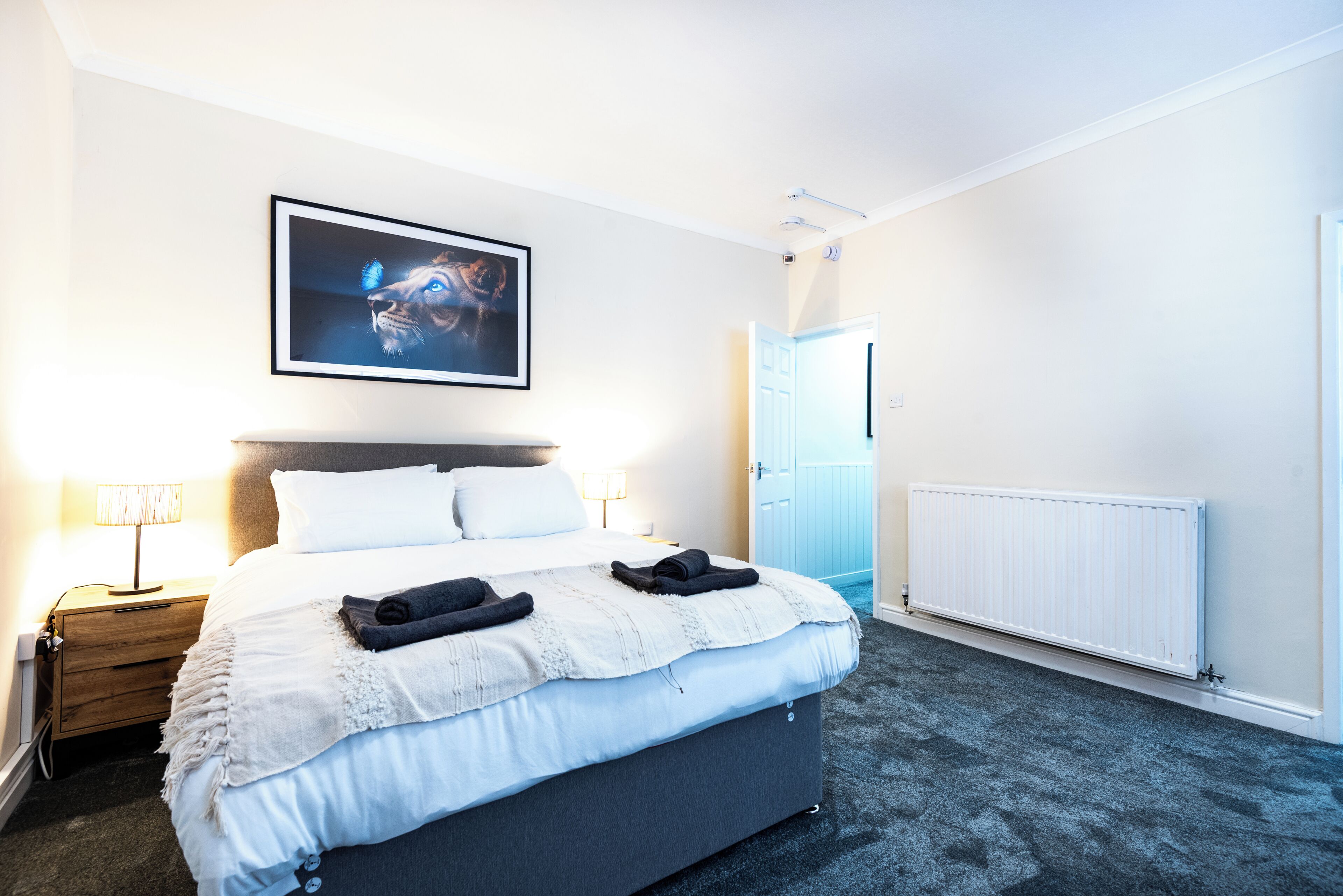 A hotel room with a bed, a nightstand, a lamp, a framed picture on the wall, and a door leading to another room.