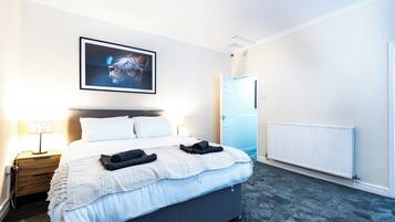 A hotel room with a bed, a nightstand, a lamp, a framed picture on the wall, and a door leading to another room.