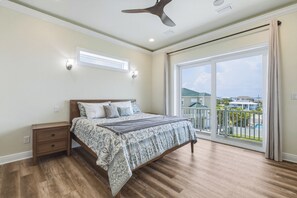 A bedroom with a large bed, a nightstand, a ceiling fan, and a view of a residential area through a sliding glass door. - Gulf Front Luxury Home | Private Pool | Rooftop Deck | Event Friendly (Pensacola Beach)