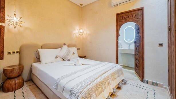 A bedroom with a bed, bedside tables, a hanging light fixture, a wall-mounted air conditioner, and a wooden door with a mirror. - Luxury Private Riad Taj 2Min Jemaa Lefna (Marrakech)