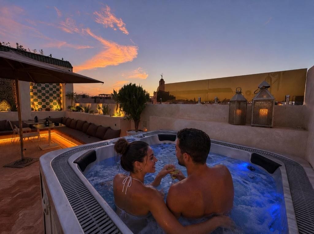 Una pareja en un jacuzzi con vista al atardecer, mobiliario exterior y faroles decorativos.