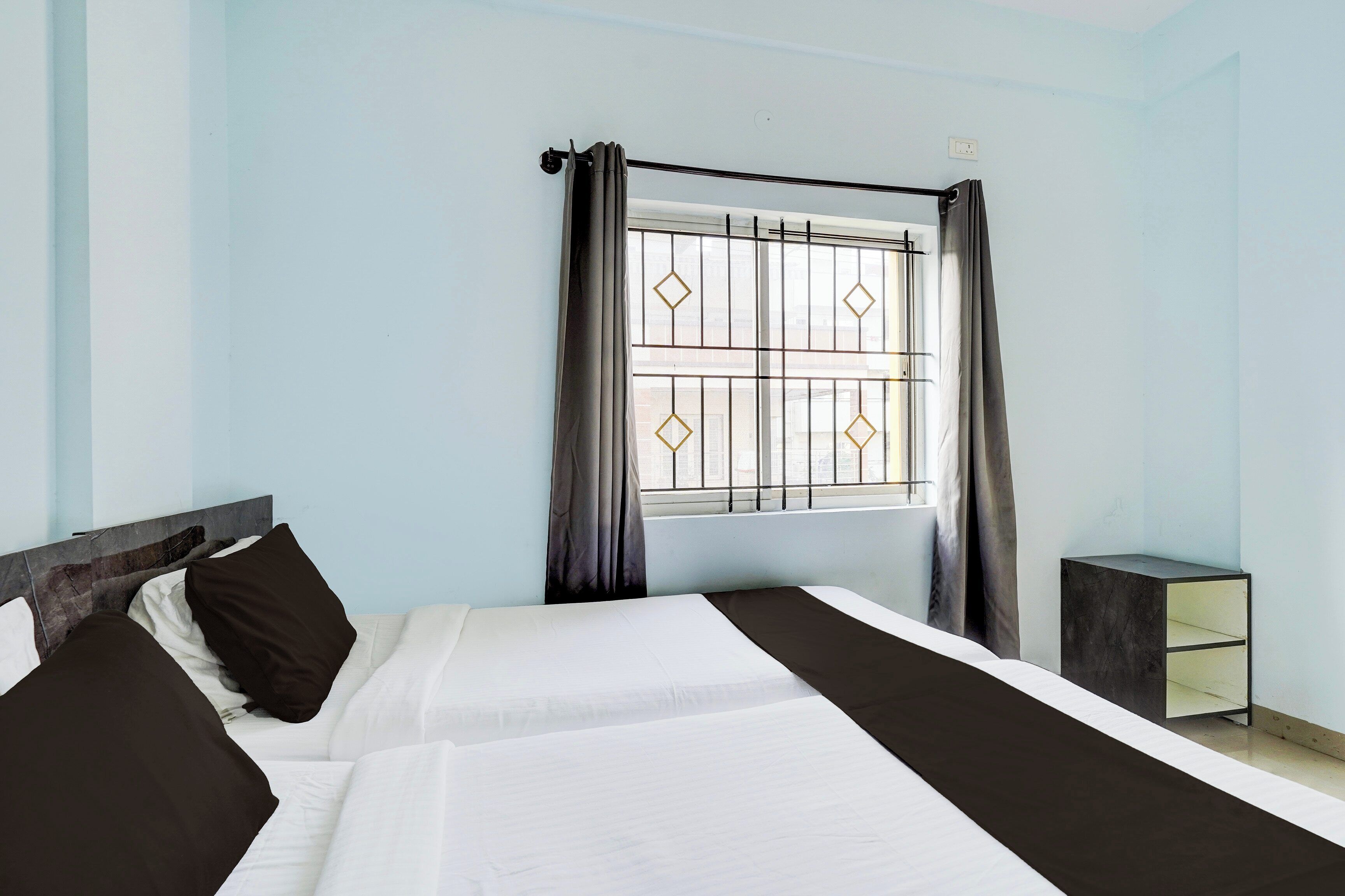A hotel room with two beds, a window with a metal grille, and a bedside table.