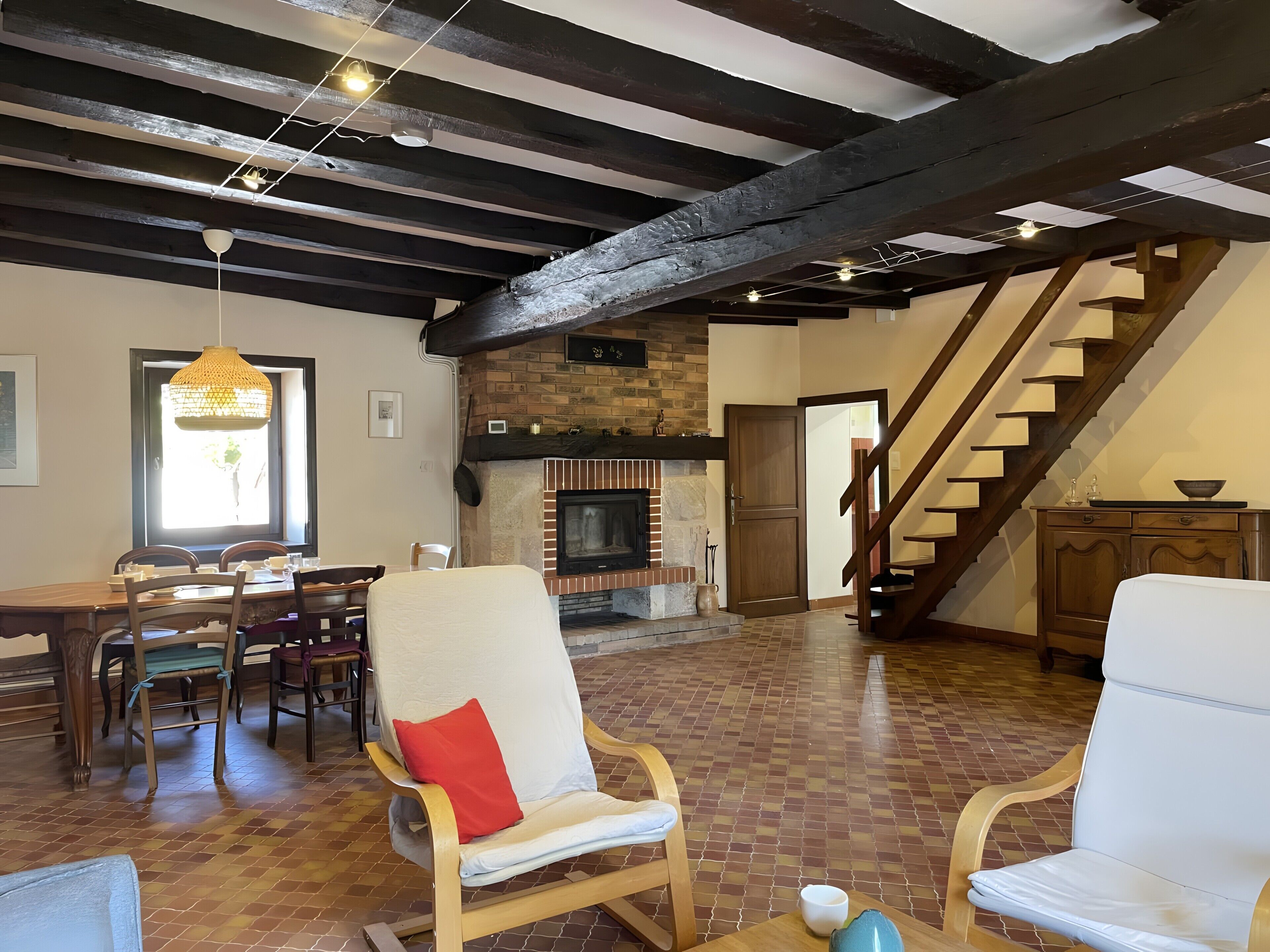A spacious living area with a fireplace, wooden beams, a dining table with chairs, and a staircase.