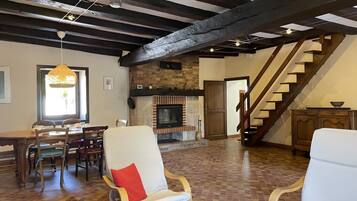 A spacious living area with a fireplace, wooden beams, a dining table with chairs, and a staircase.