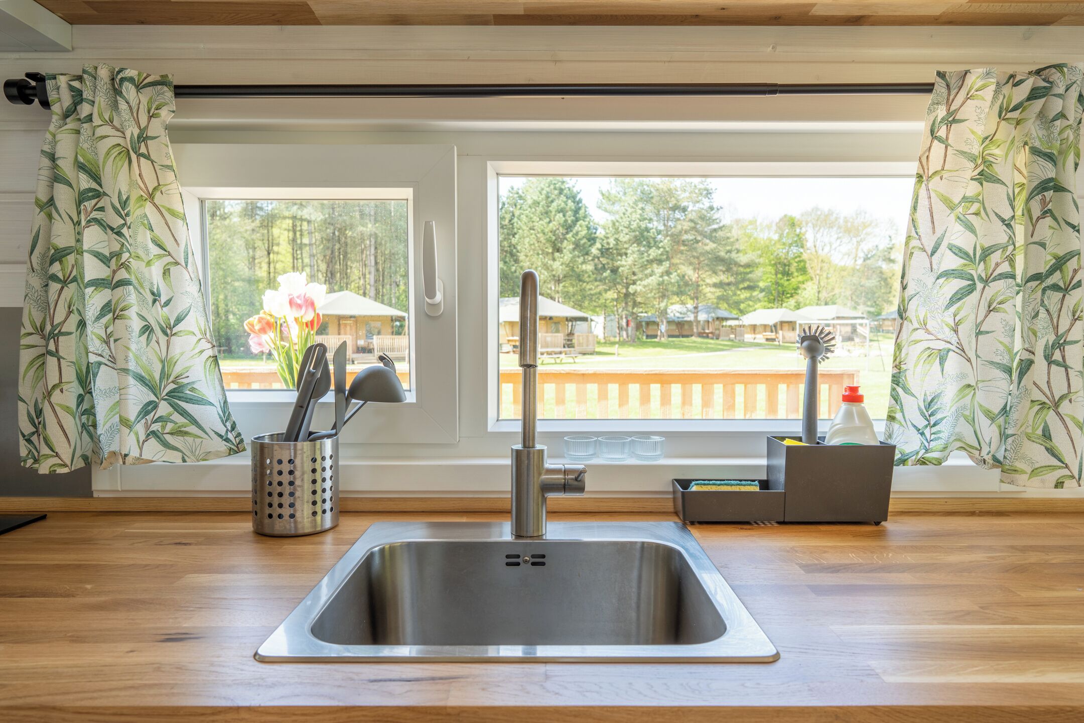 A kitchen with a stainless steel sink, wooden countertop, and a window with a view of a park and outdoor seating.