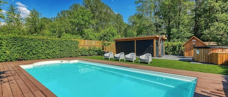 Una piscina rectangular con un deck de madera, rodeada por un área de césped y árboles. Se observan tres sillas blancas y una pequeña estructura de madera al fondo.